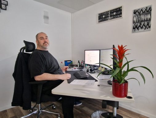 Mann sitzt an Schreibtisch, arbeitet an zwei Monitoren, Pflanze in rot-bläulichem Topf.