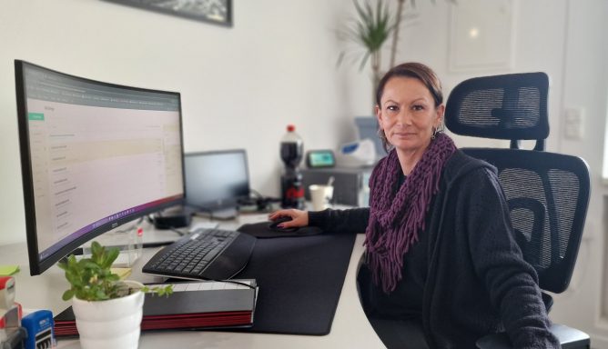 Frau sitzt an einem Schreibtisch mit Computer, Pflanzen und Büromaterialien.